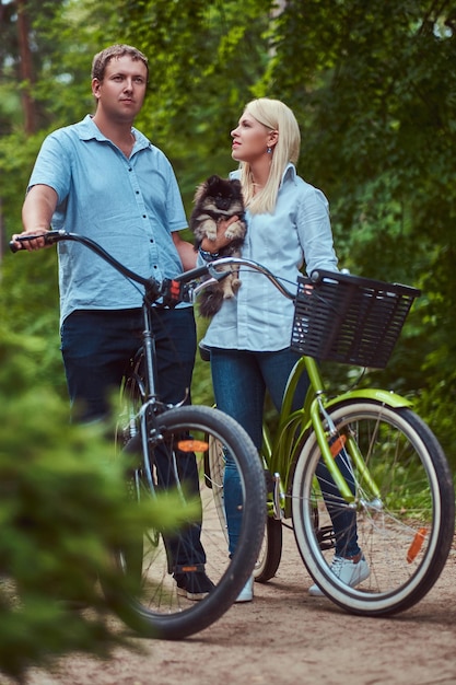 An attractive couple of a blonde female and man dressed in casual clothes on a bicycle ride with their cute little spitz.