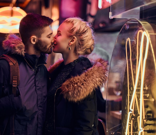 Photo attractive couple in love. a beautiful girl kisses her boyfriend standing in the night on the street. illuminated signboards, neon, lights.