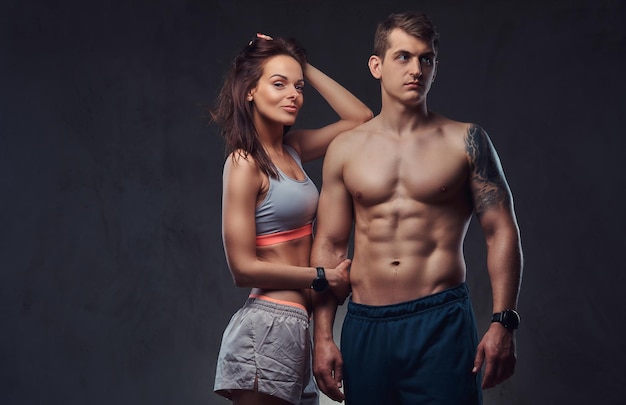 Attractive couple, a slim brunette female wearing sports bra and shorts and handsome shirtless guy cuddling in a studio. Isolated on a dark background.