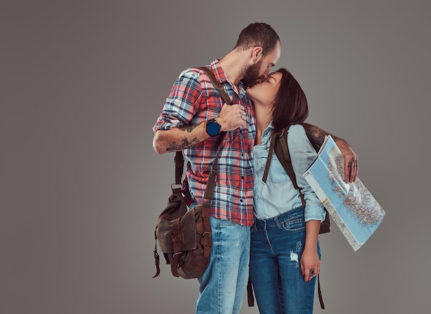 Attractive couple of tourists kissing while covering their face with a map, cuddling in a studio. Isolated on a gray background.