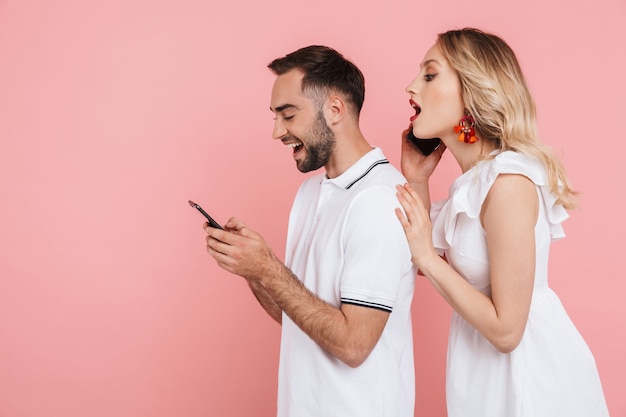 Привлекательная любопытная молодая пара, стоящая вместе изолированно над розовым, используя мобильный телефон