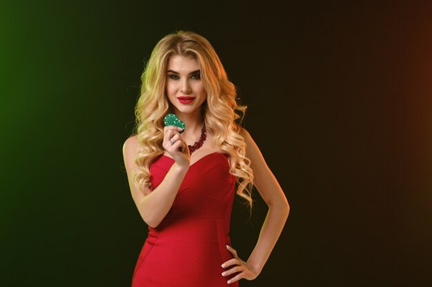 Attractive curly blonde girl, bright make-up, in red fitting dress and necklace. She smiling, showing two green chips, hand on waist, posing on colorful background. Poker, casino. Close-up, copy space