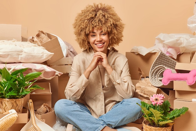 Attractive curly haired woman sits in lotus pose on floor keeps\
hands under chin smiles broadly happy to move to new apartment\
packs things in cardboard boxes isolated over beige background