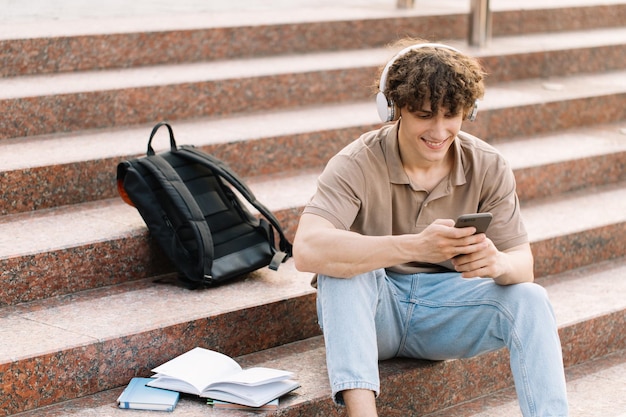 Attractive curly haired young man university or college student sits on the stairs and looking smartphone