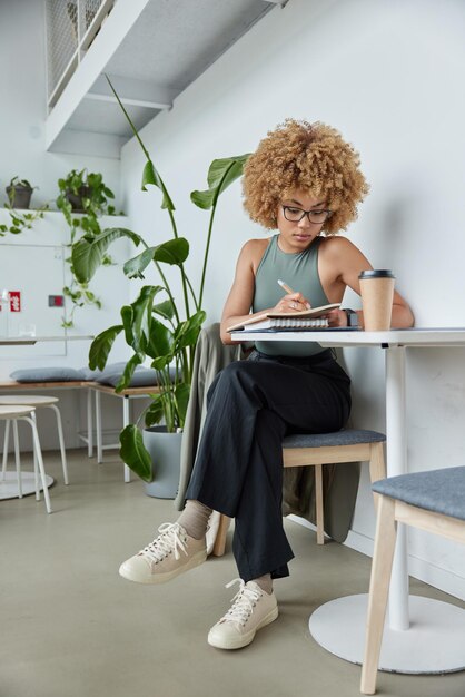 Attractive curly haired young woman writes list to do in\
notebook notes text information dressed in stylish clothes drinks\
takeaway coffee poses at cozy coffee shop student prepares for\
exams