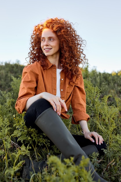 Attractive Curly Red-haired Woman Sits On Grass In Nature Countryside, Relaxing And Resting Alone, Portrait Of Female Traveler Having Leisure, Happy Weekends Outdoors, In Rural Environment