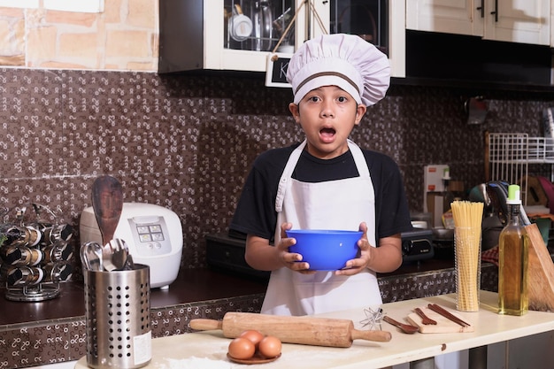 Attractive cute boy using chef uniform showing shocked expression while holding bowl of dough