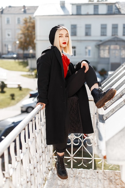 革の靴のズボンのスタイリッシュな赤いシャツの黒いコートのヴィンテージベレー帽の赤い唇を持つ魅力的なかわいい女性のブロンドは、街の屋外で休んでいます