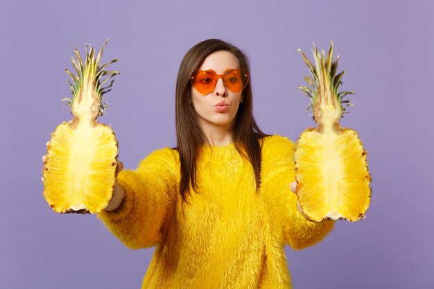 Photo attractive cute young woman in heart glasses holding in hands halfs of fresh pineapple fruit isolated on violet pastel background. people vivid lifestyle relax vacation concept. mock up copy space.