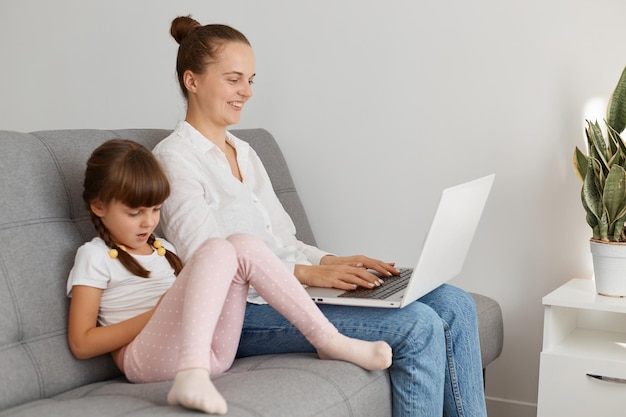 Attractive dark haired mom with hair bun and her little kid using notebook together, resting on sofa in cozy living room, making video call, watching movie, shopping online, playing games.