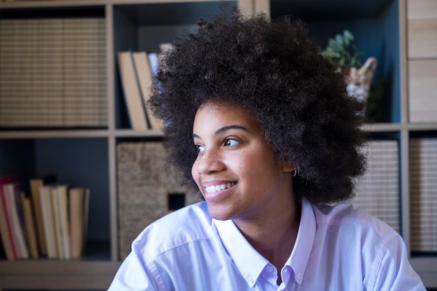 Attractive dark skinned young woman with curly Afro hairstyle looking out through window Thoughtful african american businesswoman smiling and looking away at office Female lady dreaming about future