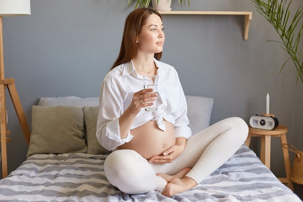 Attractive dreamy pregnant woman with bare belly wearing white clothing sitting in bed in light bedroom touching her belly and caring about her health drinking fresh water