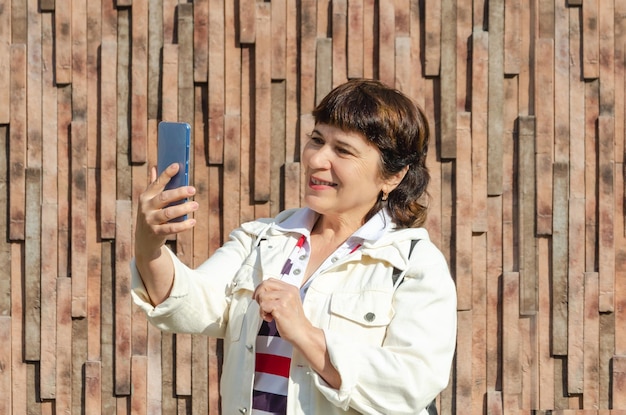 Attractive elderly woman takes selfie or video call using smartphone against background stone wall