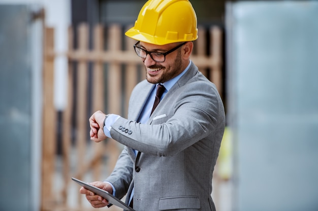 Attractive elegant caucasian smiling architect in suit, with eyeglasses and helmet on head standing at construction site and holding tablet while looking at wristwatch.