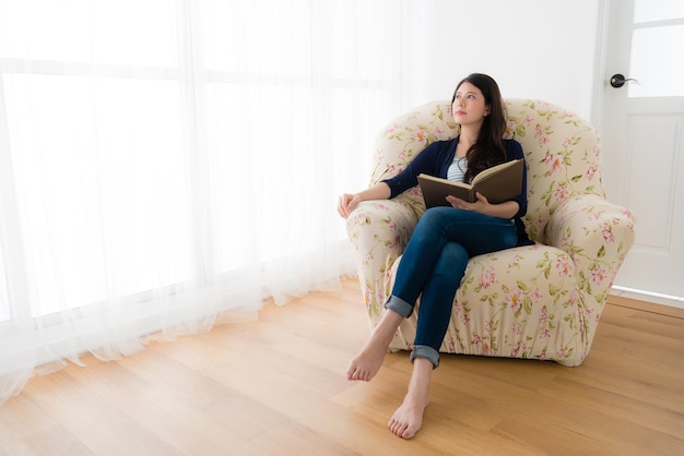 attractive elegant woman daydreaming thinking family travel planning and sitting on sofa in front of window holding dairy book looking at window at living room.