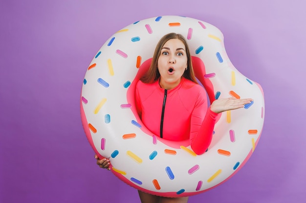 Attractive emotional woman in stylish pink swimsuit with donut inflatable ring on plain background Beach fashion