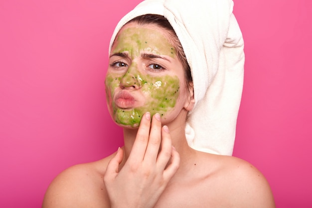 Attractive emotional young beauty has green mask on her face, day to have spa procedures, protrudes her lips, has unpleasant facial expression. Magnetic model poses isolated on pink.