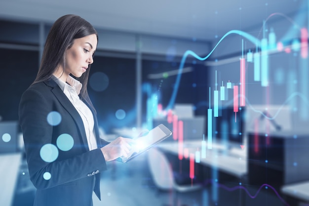 Attractive european businesswoman holding laptop with glowing business chart on blurry office interior background Trade finance market and financial growth concept Double exposure