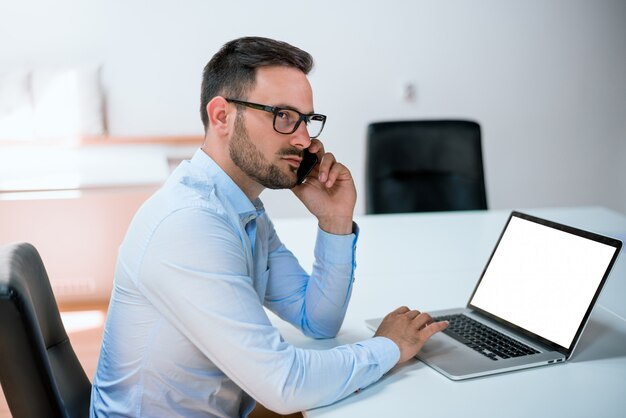 Attractive european guy talking on phone while using laptop at workplace