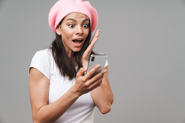 Attractive excited happy young brunette woman wearing beret standing isolated over gray wall, holding mobile phone