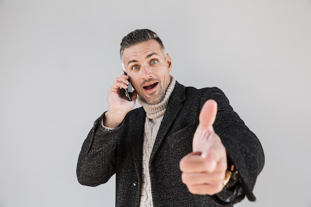 Attraente uomo eccitato che indossa un cappotto in piedi isolato su un muro grigio, parla al telefono cellulare, punta il dito davanti