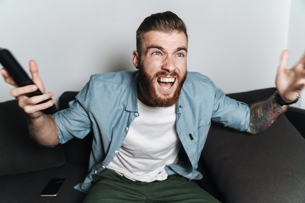 Attractive excited young bearded man relaxing on a couch at home, watching tv, screaming