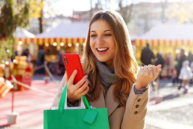 Attraente giovane donna eccitata che guarda lo smartphone e tiene in mano le borse della spesa camminando nei mercatini di natale