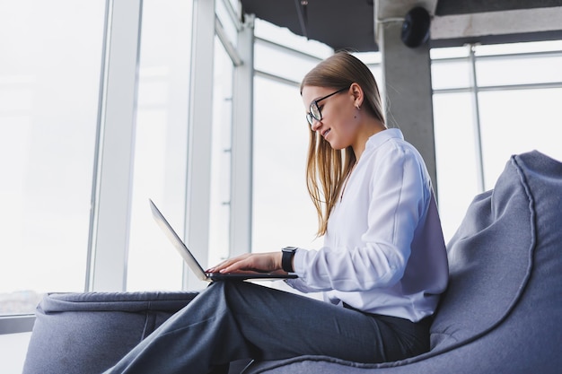 Attractive fairhaired woman in casual clothes and glasses is concentrating and typing on a laptop while sitting at a table in a cafe with coffee a student girl in a white shirt and glasses