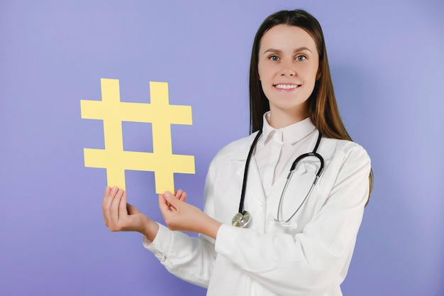 Attractive family doc lady patients consultation beaming friendly smiling reliable virology clinic holds hashtag sign wear white lab coat stethoscope, posing isolated on purple color background