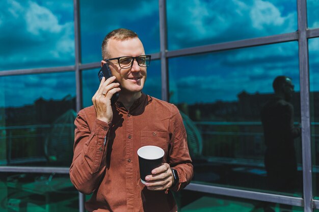 Attractive fashionable male businessman in a stylish shirt and\
trousers talking on the phone near the office building and drinking\
coffee modern glass office