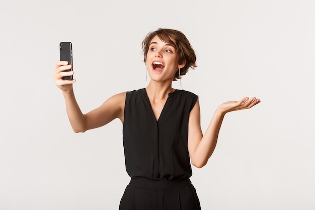 Attraente donna alla moda che ha videochiamata, dimostrando qualcosa sulla fotocamera del cellulare, in piedi bianco.