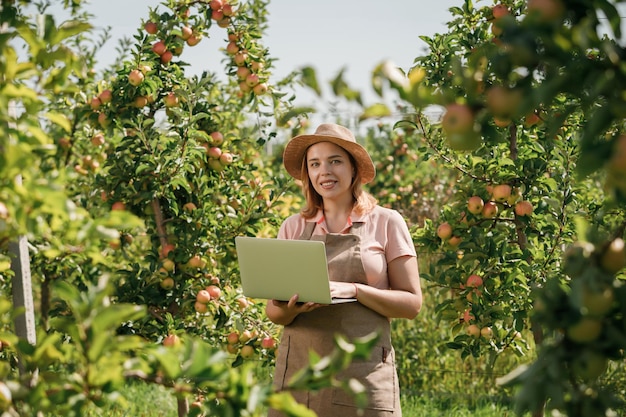 Attractive female agronomist or farmer with laptop standing in apple orchard and checking fruit makes notes Agriculture and gardening concept Healthy nutrition