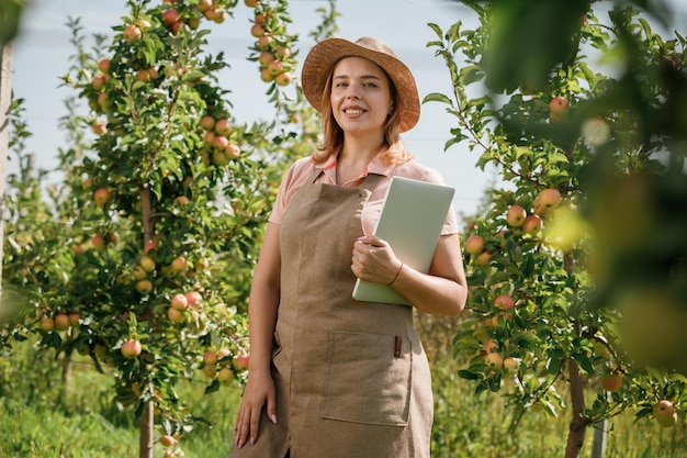 Attractive female agronomist or farmer with laptop standing in apple orchard and checking fruit makes notes Agriculture and gardening concept Healthy nutrition