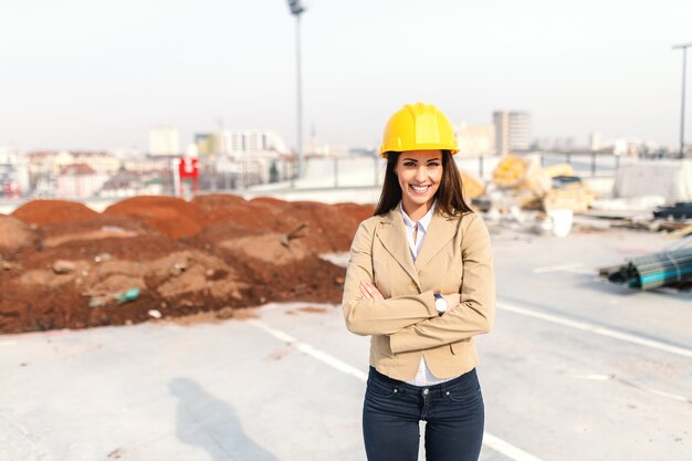 Attractive female architect with brown hair, toothy smile, helmet on head and dressed smart casual standing on construction site with arms crossed.