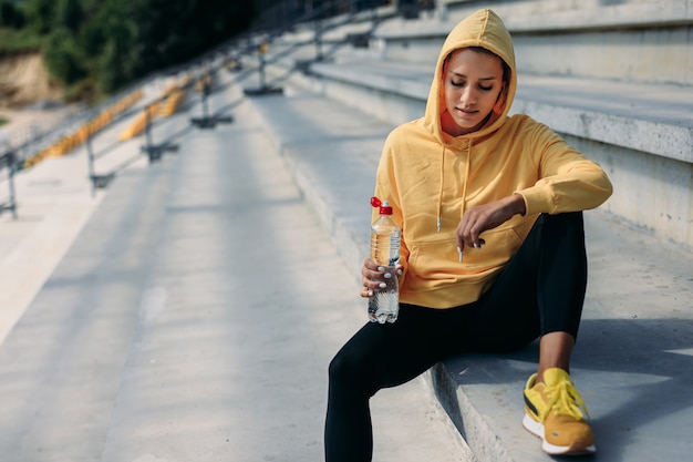 黄色いセーターの頭にフードが付いた魅力的な女性アスリートが、ボトル入り飲料水を持って階段でリラックスし、見下ろしています。