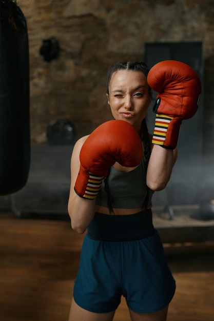 Привлекательная боксерка, глядящая через красные боксерские перчатки, с гримасом на лице. Спортивная спортсменка, гуляющая во время тренировочных упражнений в тренажерном зале