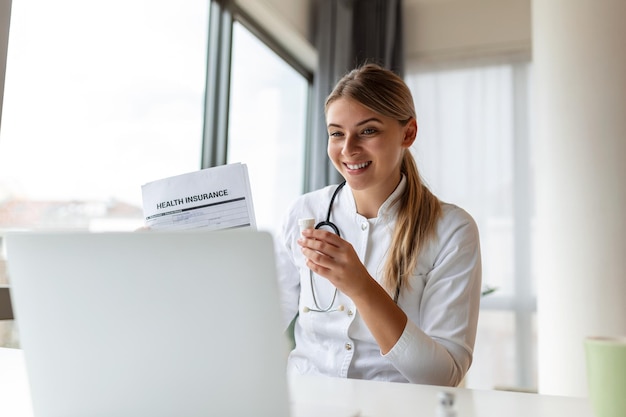 Attractive female doctor talking while explaining health\
insurance to patient through a video call with laptop in the\
consultation