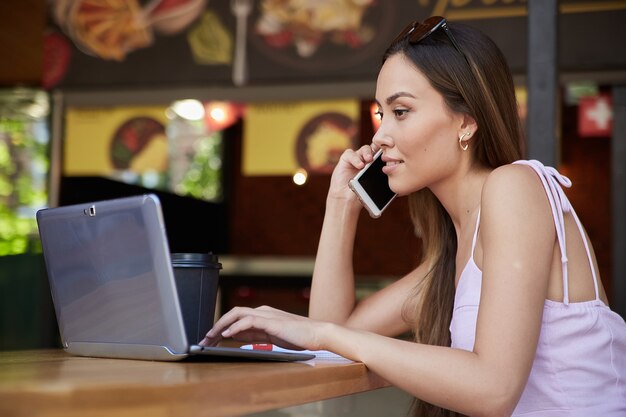 Attractive female in dress with cup of coffee working at cafe calling by phone