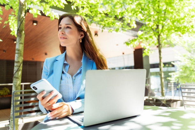 Attractive female freelancer sitting at workplace outdoors holding mobile phone using laptop