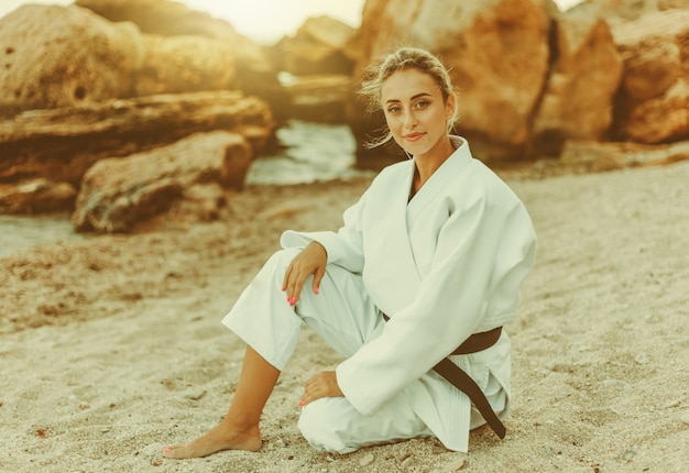 Attractive female martial artist in white kimono with black belt sits on sand wild beach