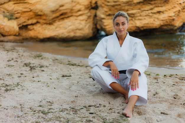 Attractive female martial artist in white kimono with black belt sits on sand wild beach