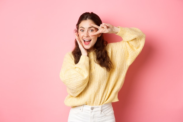 Attractive female model gasping amazed showing vsign over eye smiling happy standing against pink wall