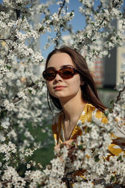 Attractive female model in sunglasses looking at camera near blooming tree with flowers in park