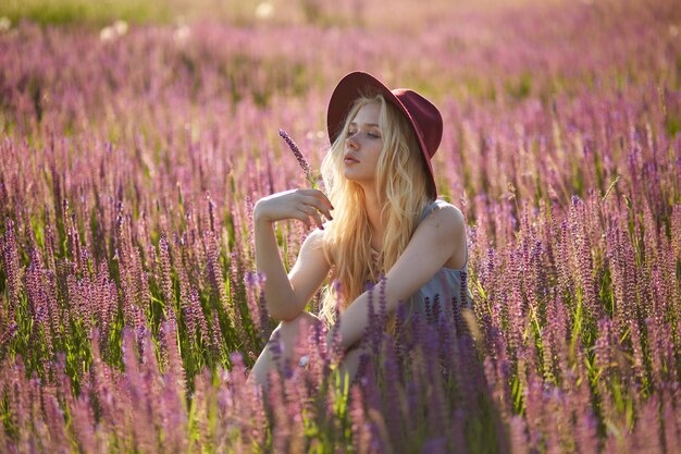 美しい夕日の背景の後ろの花畑でポーズをとるブロンドの髪の帽子をかぶった魅力的な女性モデル