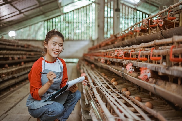 Attractive female poultry farmer smiling hold clipboard squatting with chicken farm background