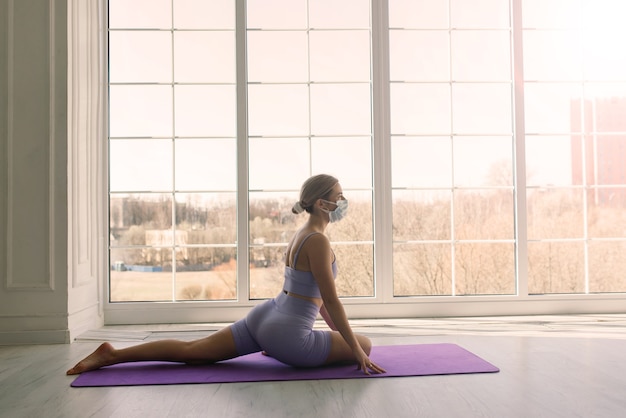 Attraente allenatore di fitness sportivo femminile che indossa una maschera medica bianca fa esercizi sul tappetino da yoga a casa.