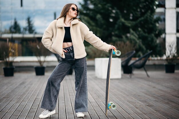 Attractive female in trendy street clothes standing with longboard on rooftop and looking aside Confident brunette wearing dark jeans beige fur coat and sunglasses