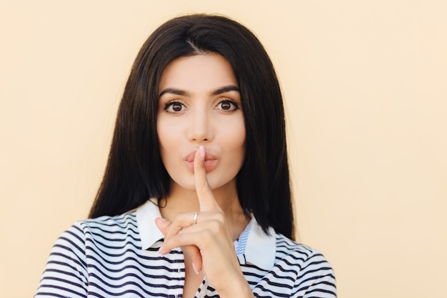 Attractive female with silence sign keeps fore finger on lips