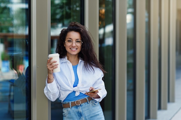 Attractive female worker with cup of tasty coffee speaks by smartphone near business centrum it