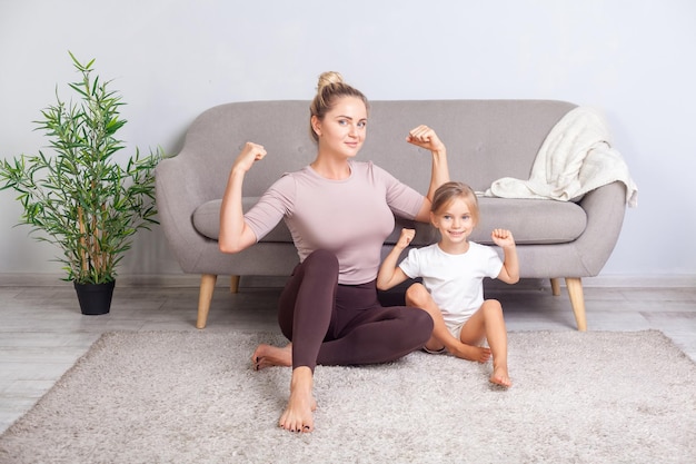 Attractive fit mom and her charming daughter showing biceps smiling at camera sitting together on floor after yoga workouts at home feeling strength and power in trained body healthcare sport